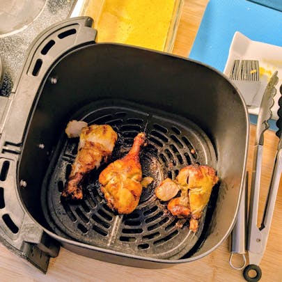 Three roasted tandoori chicken drumsticks with charred skin sitting in a black air fryer basket next to tongs and a bowl of yellow marinade.