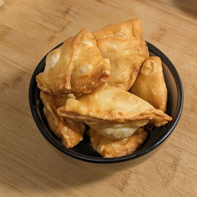 A top-down view of a small black ceramic bowl filled with several golden-brown, deep-fried samosas resting on a light-colored wooden surface.