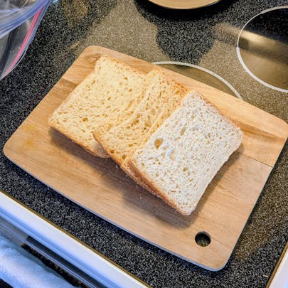 Three thick slices of airy white bread arranged on a wooden cutting board over a black stovetop.