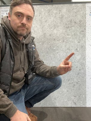 Chris standing in a bathroom panel showroom pointing at a marble effect shower wall panel .