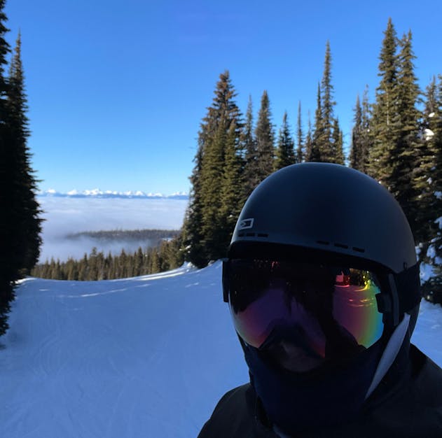 A skier wearing a helmet and colourful goggles poses on a snowy slope, surrounded by evergreen trees and a clear blue sky.