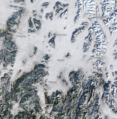 Satellite view of British Columbia, Canada, highlighting towns like Vernon, Kamloops, and Penticton among snow-covered mountains and clouds.