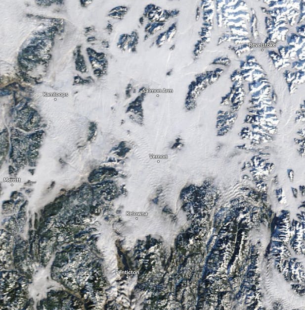 Satellite view of British Columbia, Canada, highlighting towns like Vernon, Kamloops, and Penticton among snow-covered mountains and clouds.