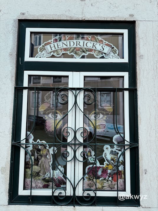 Old window with white frame and dark green border, covered by a black wrought‑iron grille; the top pane shows a ‘HENDRICK’S’ sign and the glass is filled with colorful, vintage‑style illustrations.